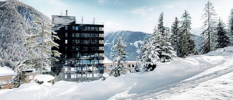 Un bâtiment moderne à plusieurs étages, entouré d’arbres et de montagnes couverts de neige.