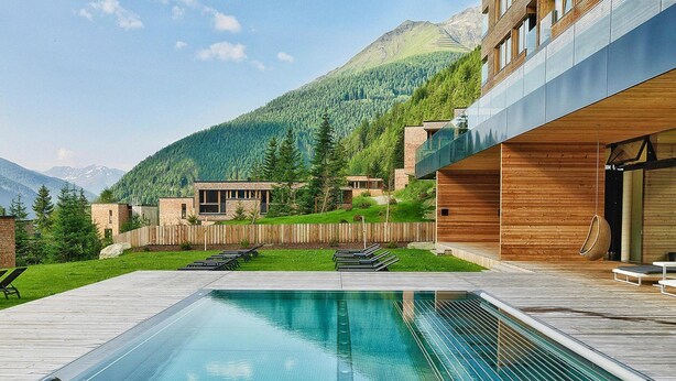 A swimming pool with wooden deck and lounge chairs, surrounded by a wooden fence and a mountain backdrop. - Chalet Resort in Kals (Kals am Grossglockner)