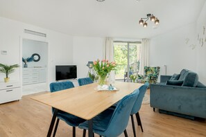 A modern living room with a wooden dining table, blue upholstered chairs, a blue sofa, and a television. - Beachfront Apartment With Indoor Pool and Sauna (Rewal)