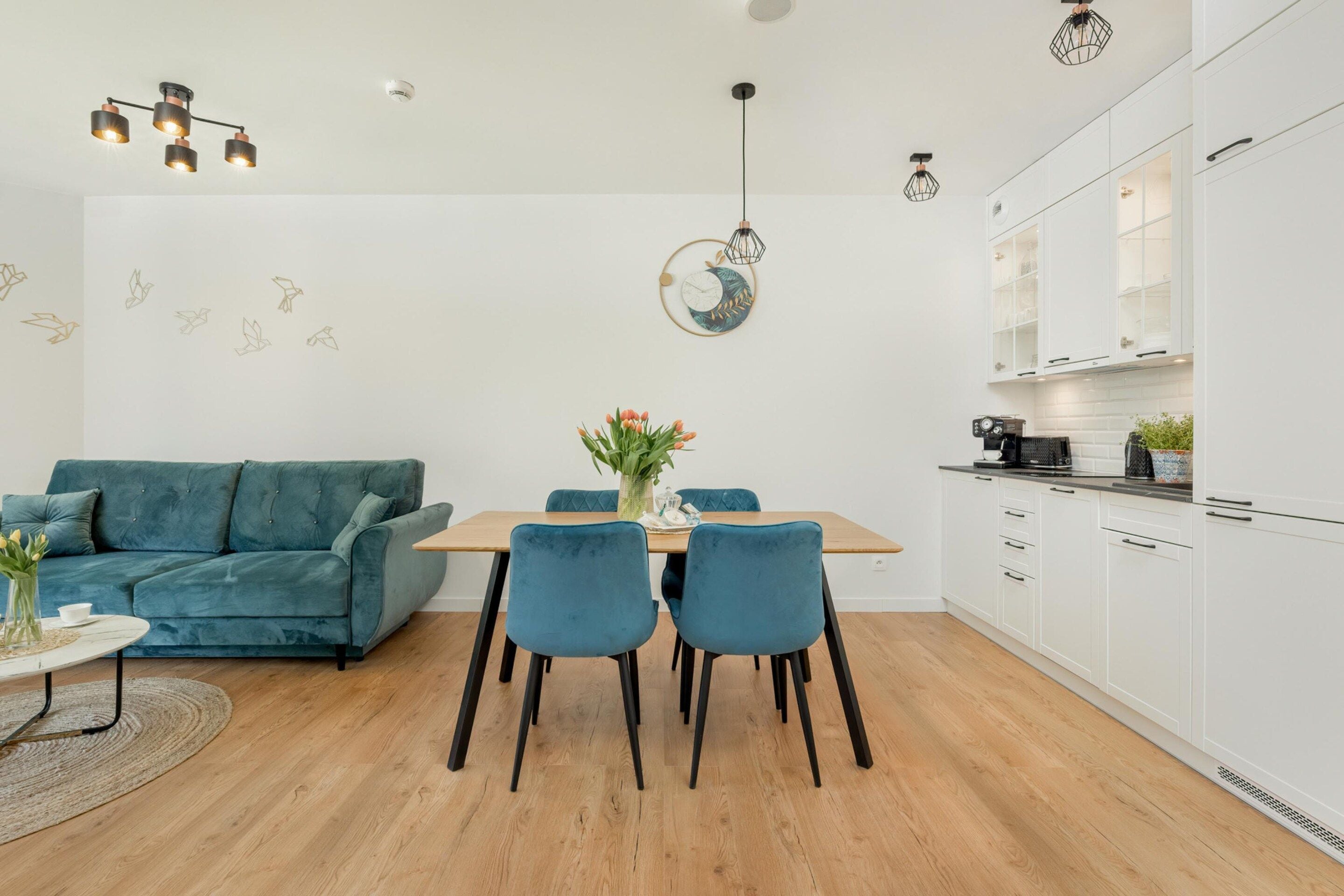 Un espace de vie moderne avec un canapé de couleur turquoise, une table à manger en bois avec des chaises, et un coin cuisine doté de meubles blancs et d’une machine à café.
