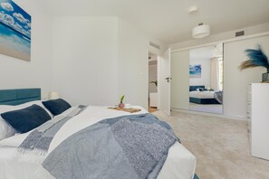 A modern bedroom with a large bed, a bedside table, a nightstand, a wall-mounted lamp, and a door leading to another room. - Beachfront Apartment With Indoor Pool and Sauna (Rewal)