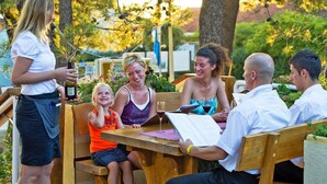 Een groep mensen zit aan een houten tafel buiten, terwijl een vrouw naast hen staat met een fles in de hand.