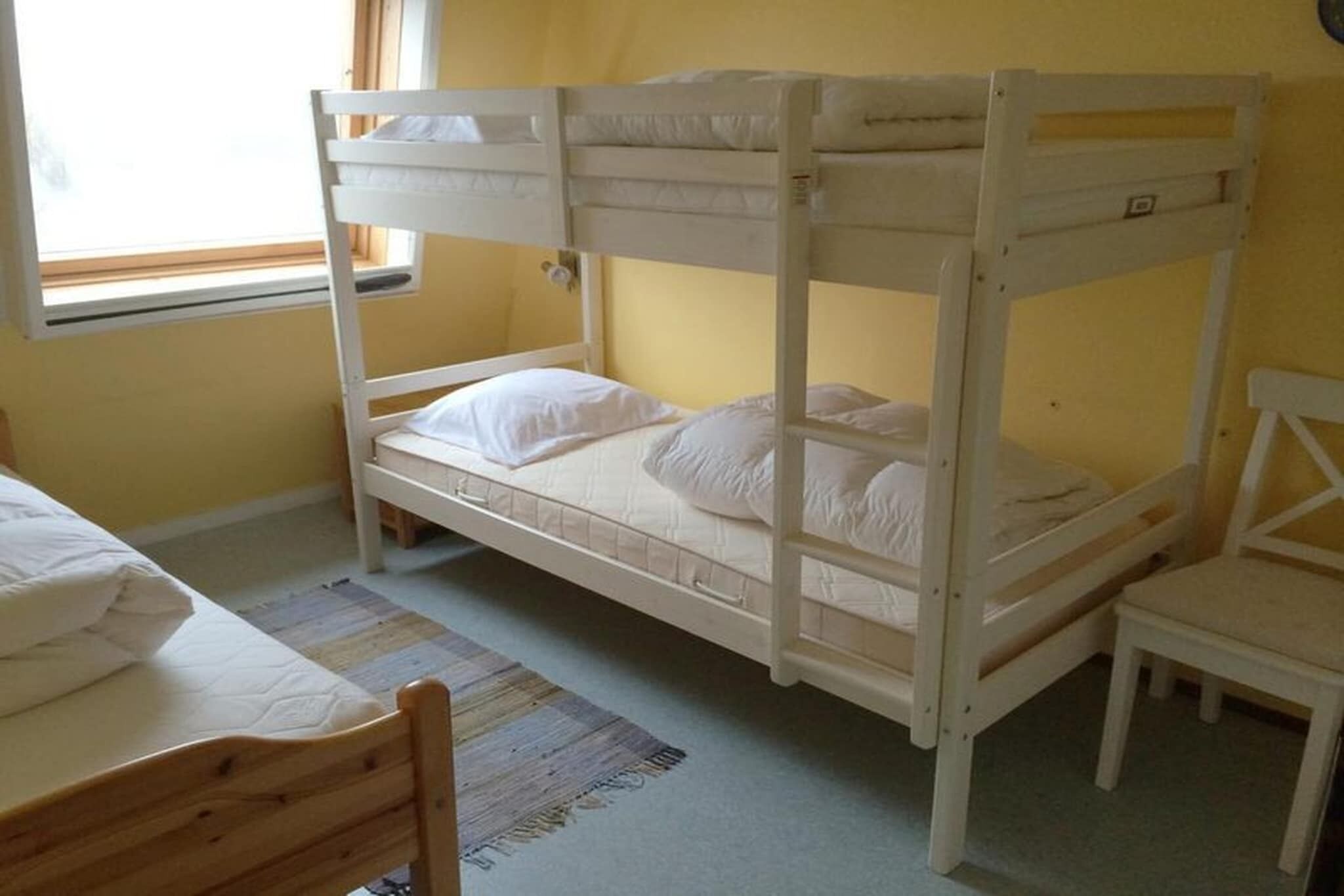 A bunk bed with a wooden frame and white bedding, a small wooden chair, and a window with curtains.