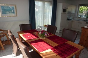 Un coin repas avec une table en bois, une nappe rouge et des chaises. On aperçoit une fenêtre avec des rideaux, une cuisine au fond et un tableau encadré au mur.