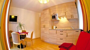 Cocina compacta con armarios de madera, un microondas y un lavamanos. Hay una zona de comedor con una mesa, sillas y una planta en maceta. Un sofá rojo es visible en primer plano.