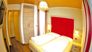 Cama doble con ropa de cama blanca, cabecera roja y lámparas de noche. Se ve un armario de madera y una puerta con un letrero.
