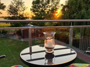 A round glass table with a candle in a glass holder, set on a balcony with a view of trees and a sunset. - Ferienhaus Weiter Feldblick auf Fehmarn (Fehmarn)