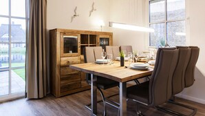 A dining area with a wooden table set for two, a cabinet with glass doors, and a view of the outdoors through large windows. - House by Bodden Lagoon With Sauna (Fuhlendorf)