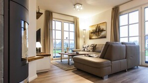 A modern living room with a sofa, a coffee table, and a TV. There are large windows with curtains, a painting on the wall, and a lamp. - House by Bodden Lagoon With Sauna (Fuhlendorf)