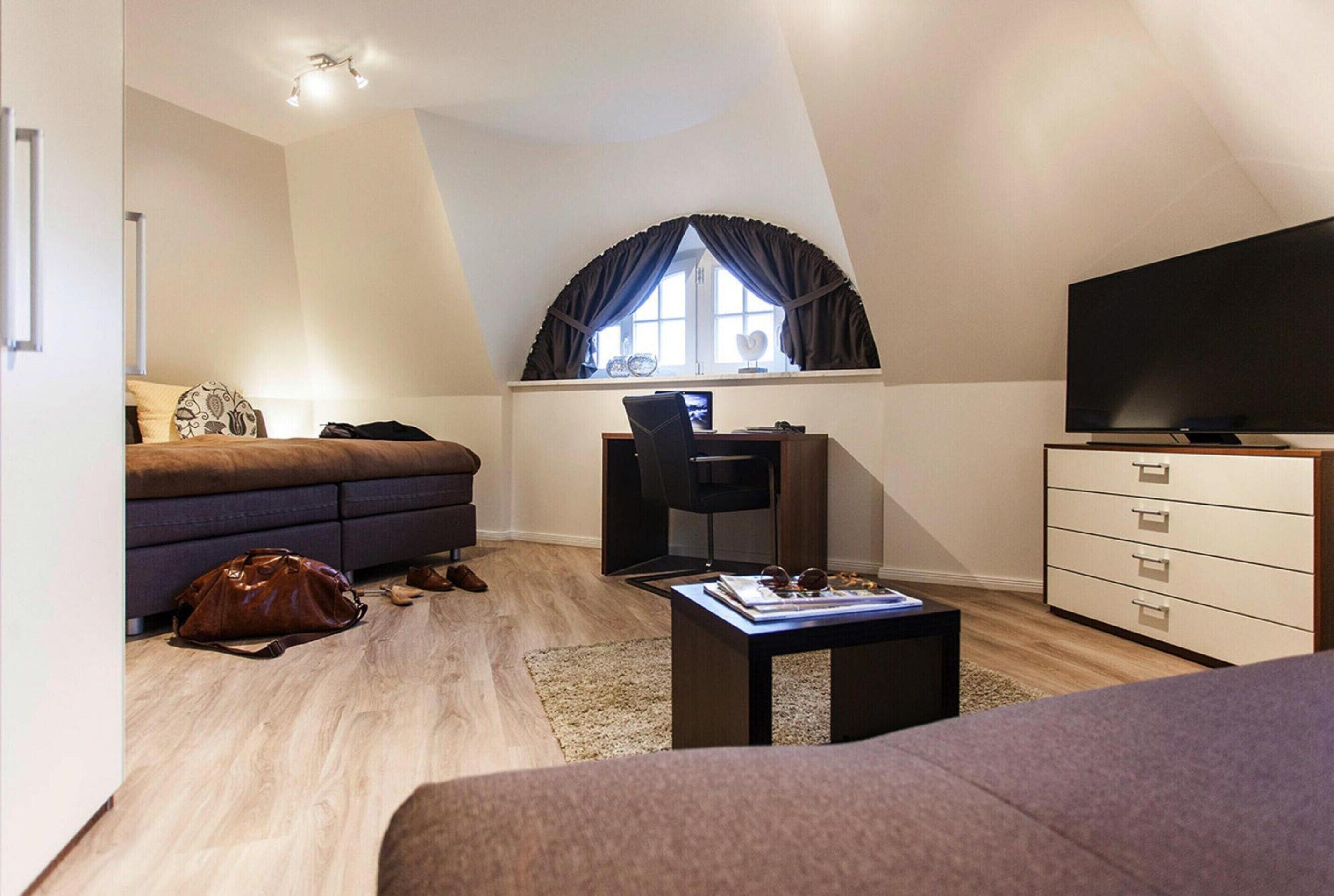 A compact, modern room with a loft-style ceiling, a sofa, a desk with a chair, a television, and a dresser.