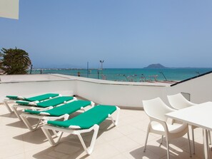 Une terrasse sur le toit, dotée de chaises longues et d’une table, offrant une vue sur une plage avec des bateaux et une montagne au loin.