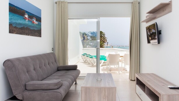 Ein modernes Wohnzimmer mit einem grauen Sofa, einem Holztisch und einem Fernseher auf einem Holzschrank. Durch die Glastüren ist ein Balkon mit Blick auf das Meer und ein Boot sichtbar.