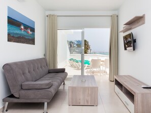 Ένα μοντέρνο σαλόνι με έναν γκρι καναπέ, ένα ξύλινο τραπεζάκι σαλονιού και μια τηλεόραση τοποθετημένη σε μια ξύλινη βάση. Μέσα από τις γυάλινες πόρτες φαίνεται ένα μπαλκόνι με θέα στη θάλασσα και ένα σκάφος.