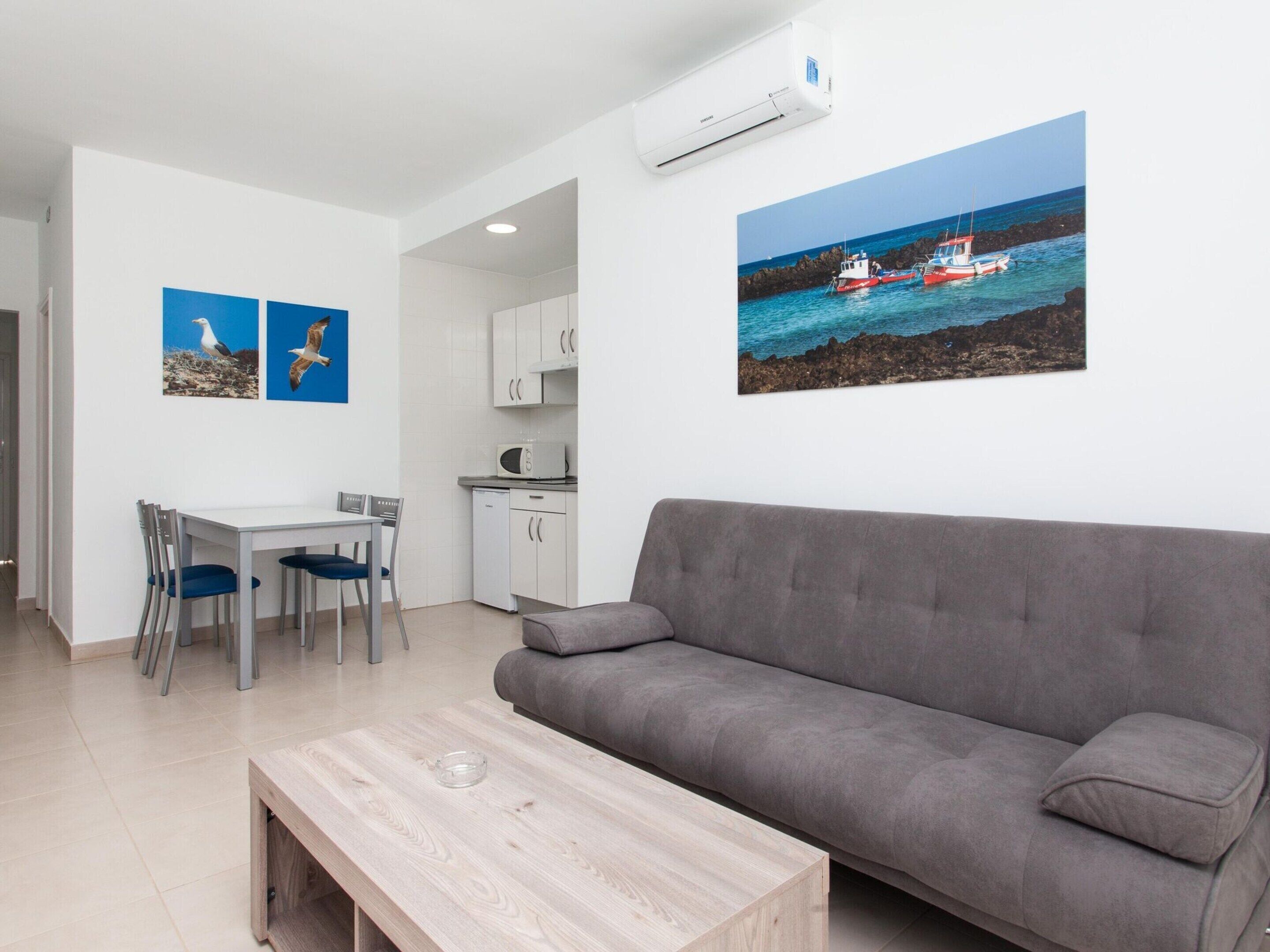 Un salon moderne avec un canapé gris, une table basse en bois et un coin repas comprenant une table et des chaises blanches. On y trouve également une cuisine avec des meubles blancs et un four à micro-ondes. Deux tableaux encadrés représentant des oiseaux sont accrochés au mur, et un grand tableau représentant un bateau sur une côte rocheuse est également visible.