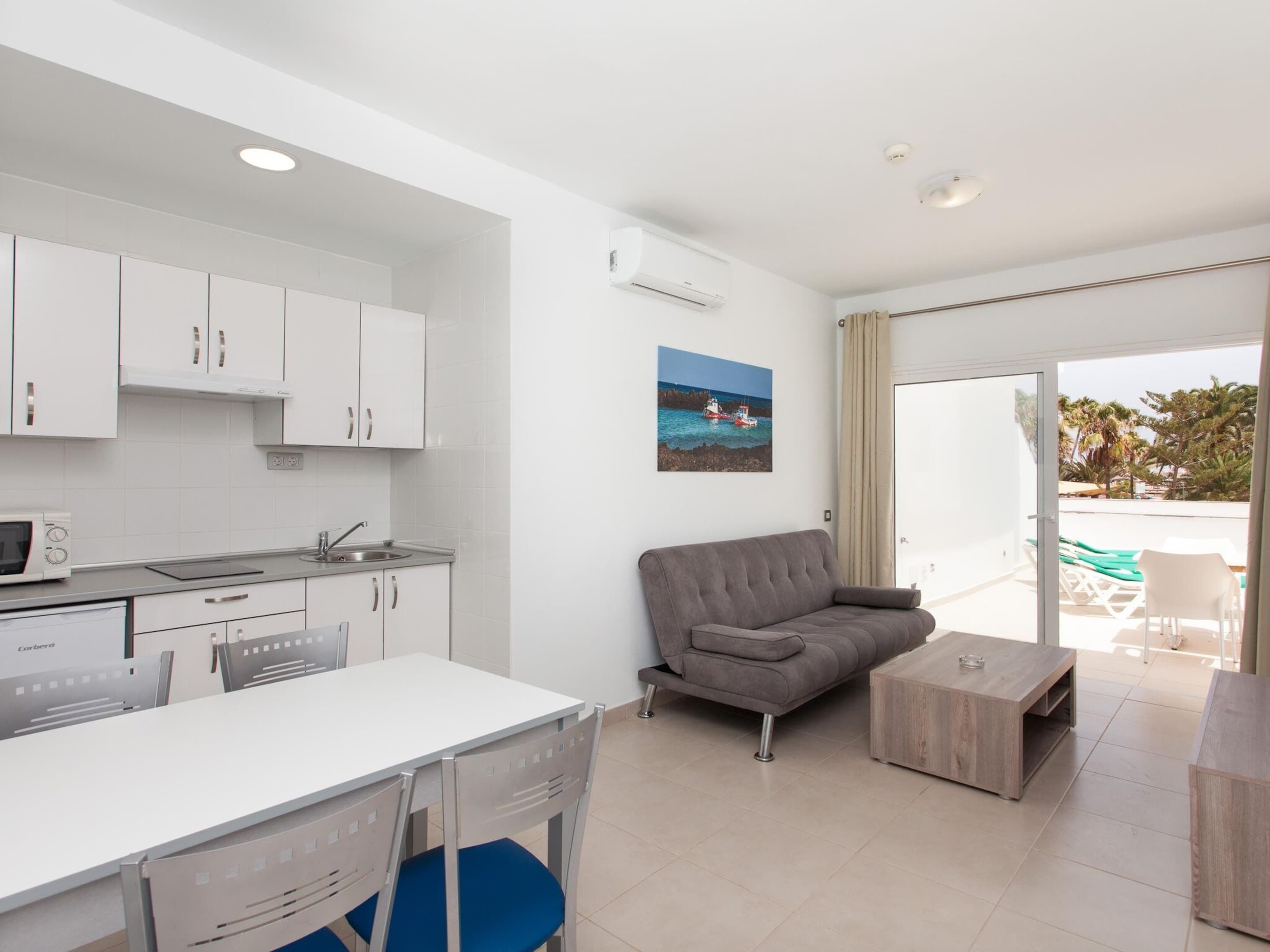 A modern kitchen with white cabinets, a dining area with a white table and chairs, a gray sofa, and a small wooden coffee table.