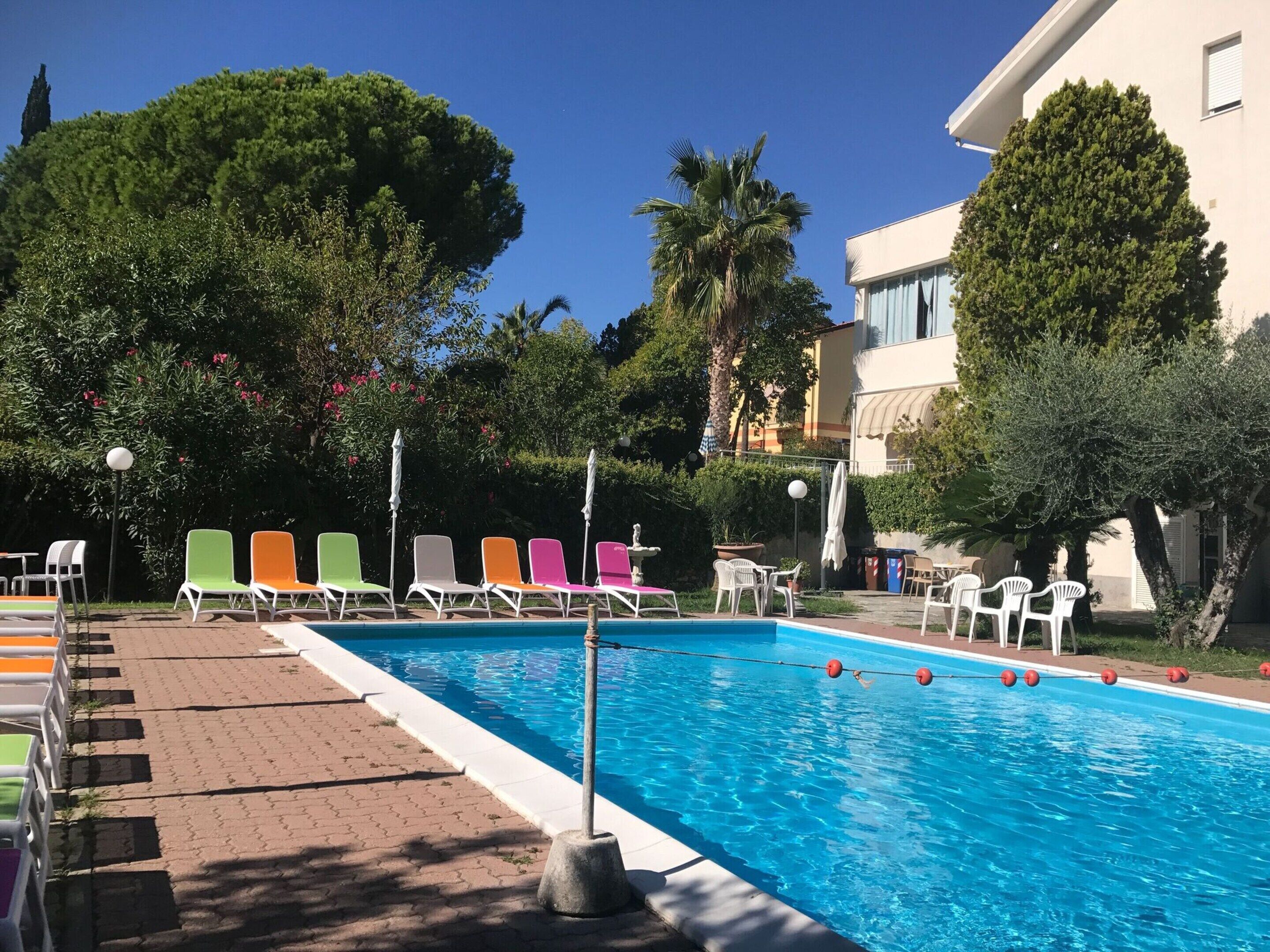 A swimming pool with colorful lounge chairs and umbrellas, surrounded by trees and a building.