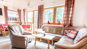 A cozy living room with a sofa, armchair, and coffee table. There are large windows with curtains, a dining area with a table and chairs, and a clock on the wall. - Chalet Hochkrimml With Sauna on the ski Slope (Krimml)