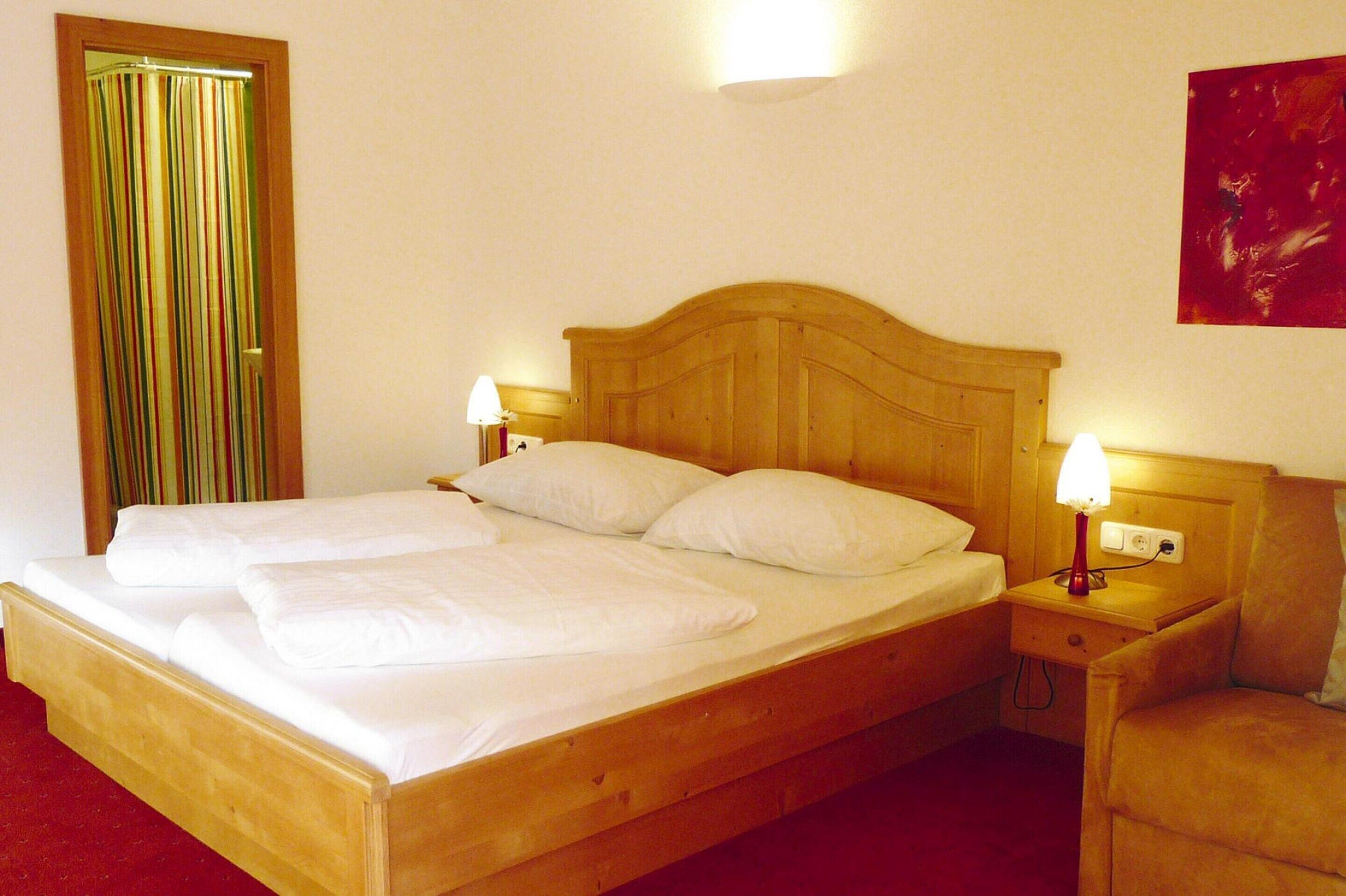 Una cama bien hecha con sábanas blancas, cabecera de madera, dos lámparas de noche, una mesita de noche y una cortina roja.