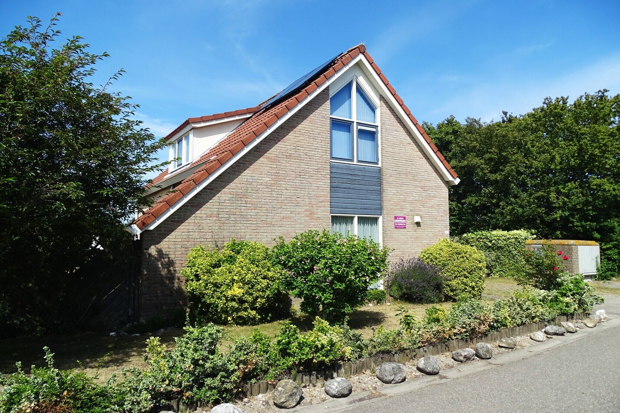 A two-story building with a sloped roof, a visible window, and a small garden area with shrubs and a pathway leading to the entrance.