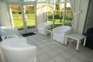 A conservatory with white furniture, a small table, and a view of a green landscape. - Bungalow in Breskens near Beach and Lighthouse (Breskens)