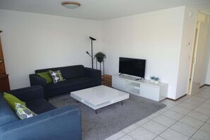 A living room with a sofa, a coffee table, a TV on a stand, and a door. - Bungalow in Breskens near Beach and Lighthouse (Breskens)
