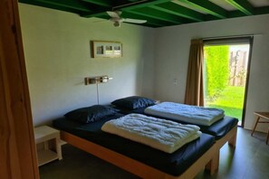 A bedroom with two beds, a ceiling fan, a small bedside table, a window with a view of greenery, and a picture on the wall.