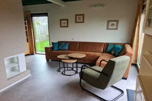 A living room with a brown sofa, two round wooden tables, a green armchair, and a sliding glass door leading to a garden.