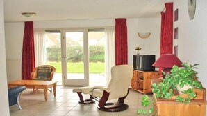 A living room with a sofa, armchair, TV stand, and a sliding glass door leading to a garden. - Waterfront Bungalow Near Beach (Breskens)