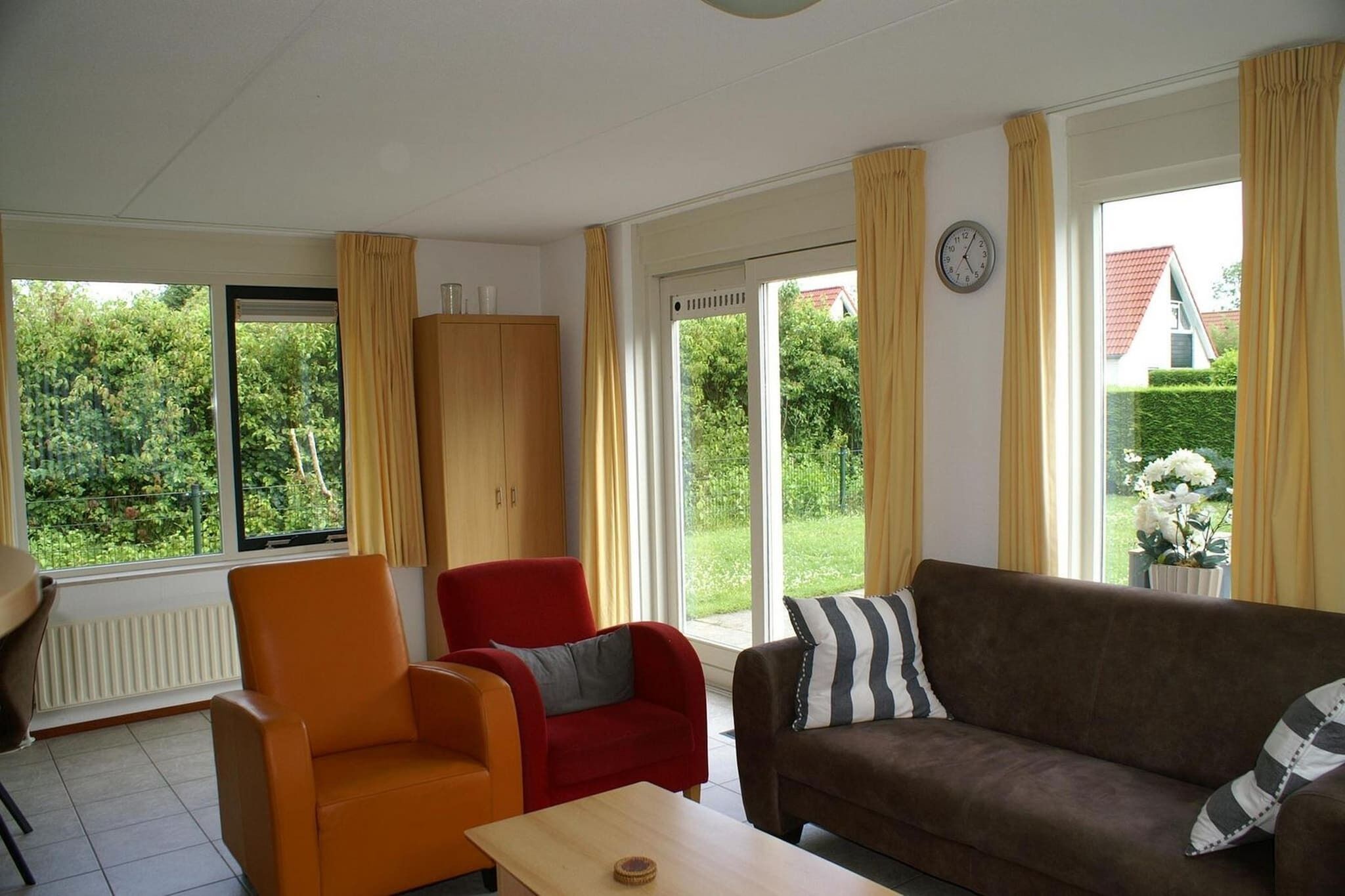 Ein Wohnzimmer mit einer Couch, Sesseln, einem Tisch und großen Fenstern mit Vorhängen.