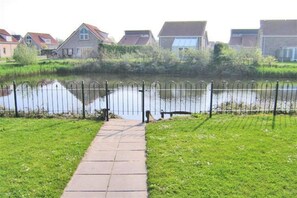 A pathway leading to a body of water with a metal fence, surrounded by greenery and residential buildings.
