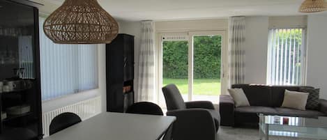 Una sala de estar moderna con una mesa de comedor, sofá y sillas, ventanas amplias con cortinas, y una lámpara colgante de mimbre.