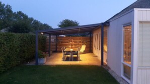 Un patio cubierto con una mesa y sillas, una cerca de madera y un edificio blanco con ventanas.