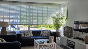 Una sala de estar con un sofá, una mesa de centro, un televisor y ventanas grandes con persianas.