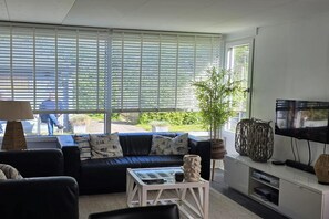 A living room with a sofa, a coffee table, a TV, and large windows with blinds.