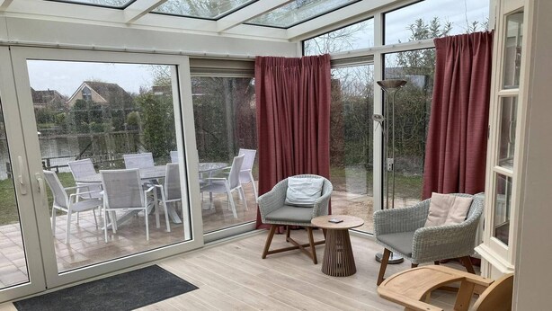 A conservatory with a glass door, a dining table with chairs, a cushioned chair, a small round table, and a wooden chair. - Stylish Coastal Home w/ Jetty (Breskens)