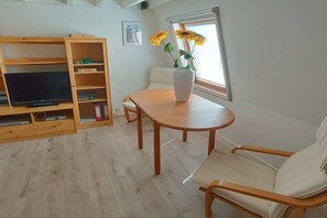 A room with a wooden TV stand, a dining table, a sofa, and a window with flowers.