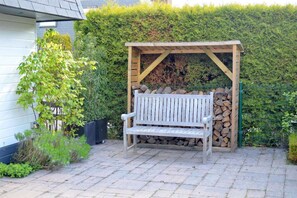 A wooden bench under a pergola with a stack of firewood behind it, set against a hedge and a house.