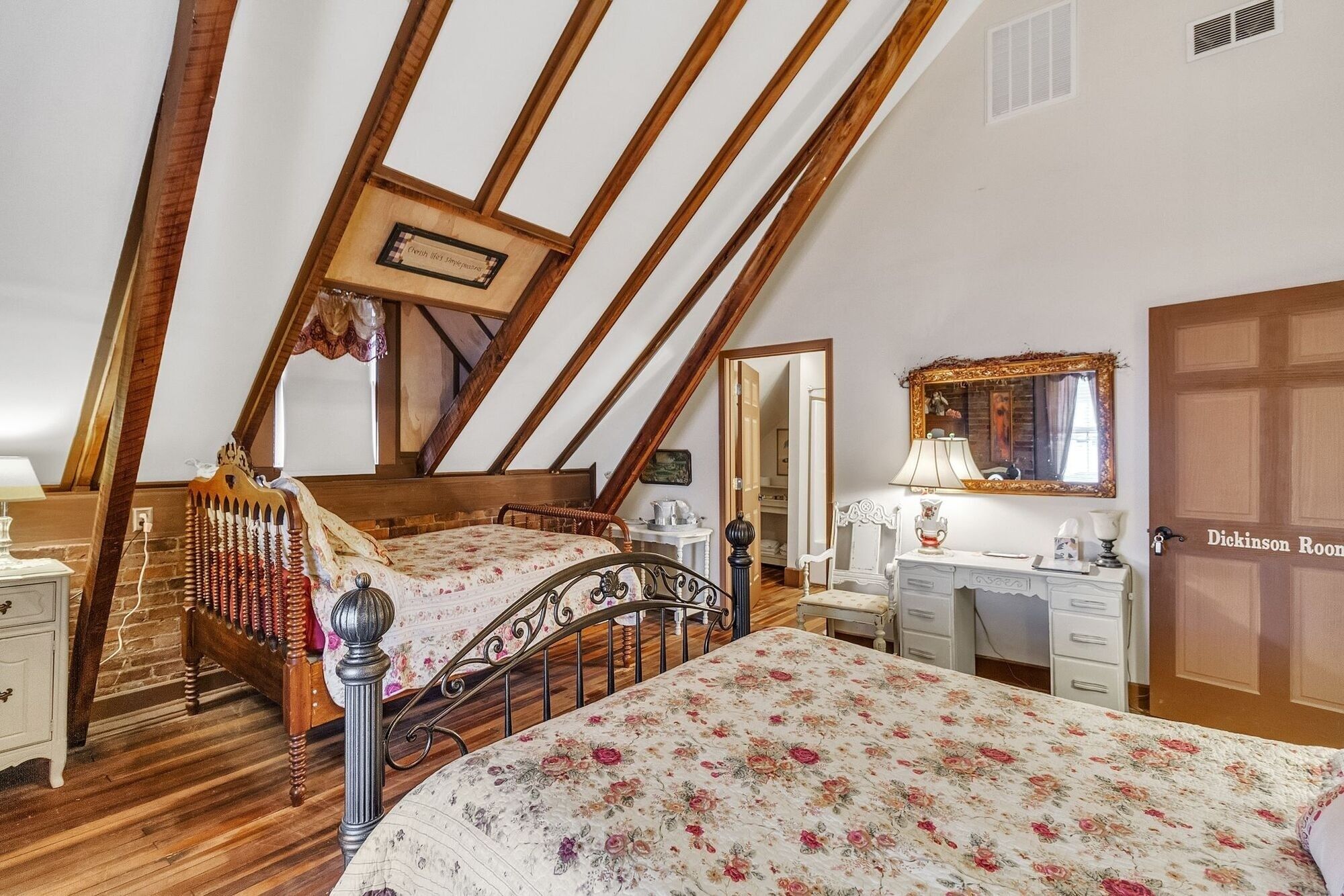 A bedroom with a sloped ceiling, a wooden bed, a bedside table, a dresser, a mirror, and a door labeled "Dickinson Room".