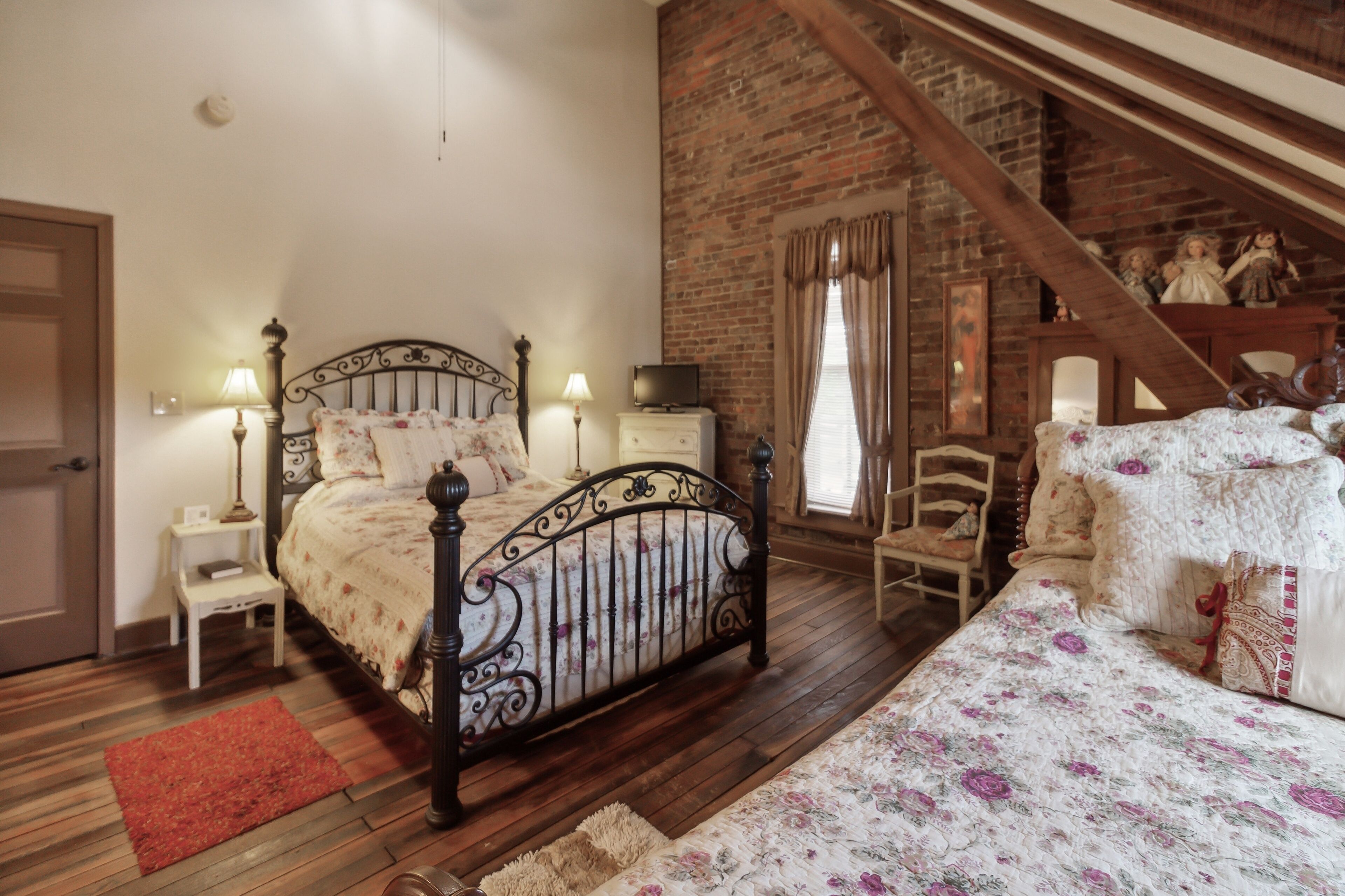 Quarto com piso de madeira, cama com colcha florida, mesa de cabeceira com abajur e parede de tijolos.