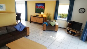 A living room with a sofa, a coffee table, a TV on a stand, a cabinet, and a painting on the wall. - Bungalow in Breskens near the Lighthouse (Breskens)