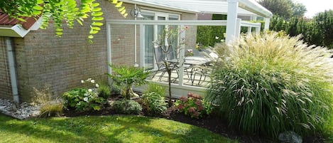 A conservatory with a glass roof, a brick house, and a garden with various plants and a lawn.