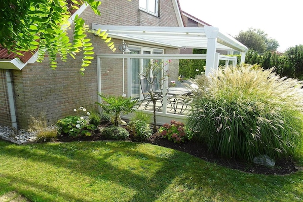 A conservatory with a glass roof, a brick house, and a garden with various plants and a lawn.