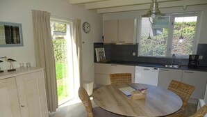 Une cuisine avec une table à manger, des chaises en osier, une horloge et une fenêtre avec des stores.