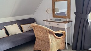 Ein Zimmer mit grauem Sofa, einem Korbstuhl, einer weißen Kommode mit Spiegel und einem kleinen Tisch mit Lampe.