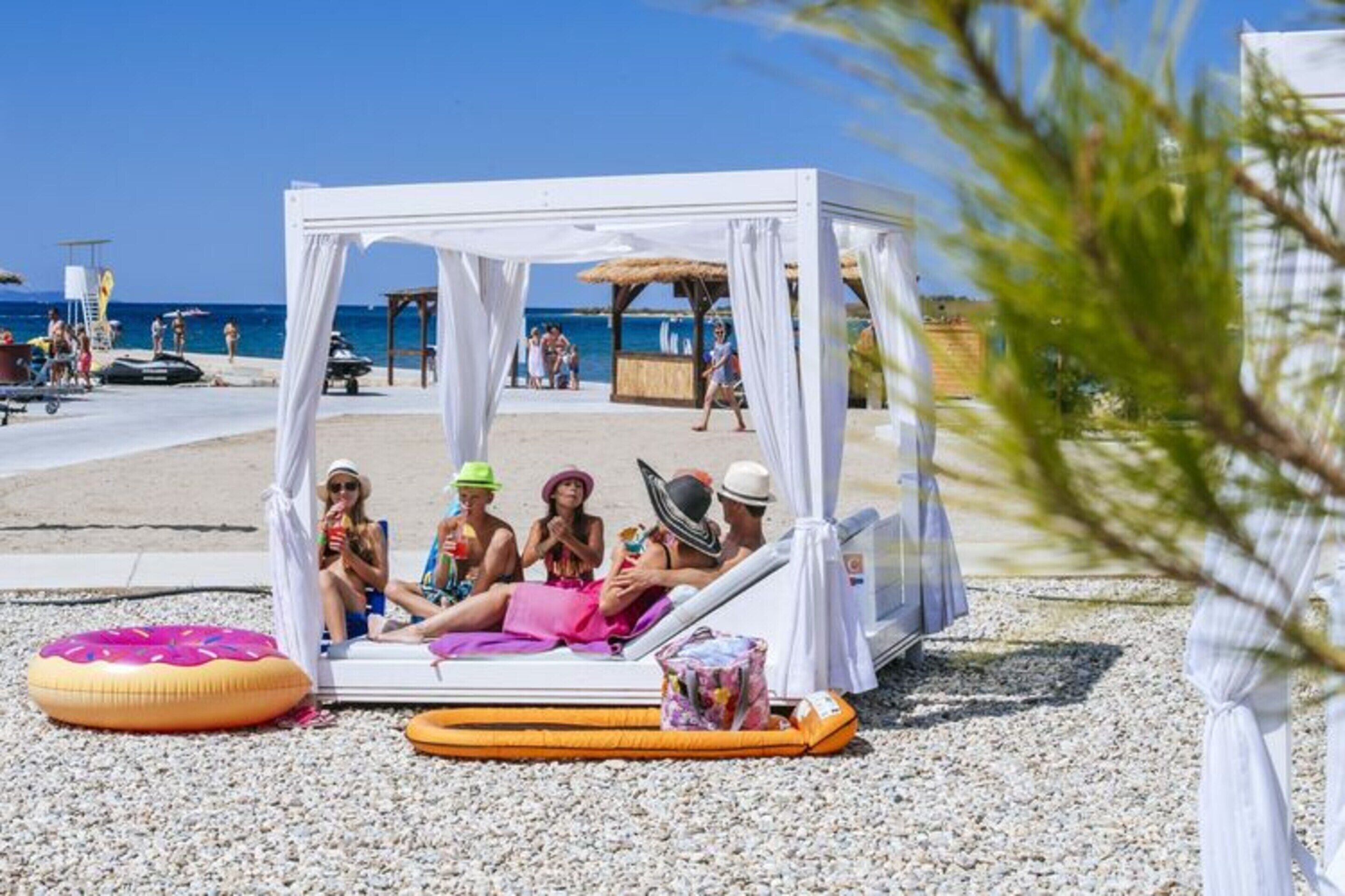 Una cabaña en la playa con toldo blanco, rodeada de guijarros, y personas relajándose en su interior.