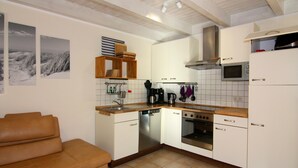 Cocina compacta con armarios blancos, lavavajillas, estufa y microondas. Hay una repisa de madera sobre el fregadero y un sofá de cuero marrón en la sala de estar.