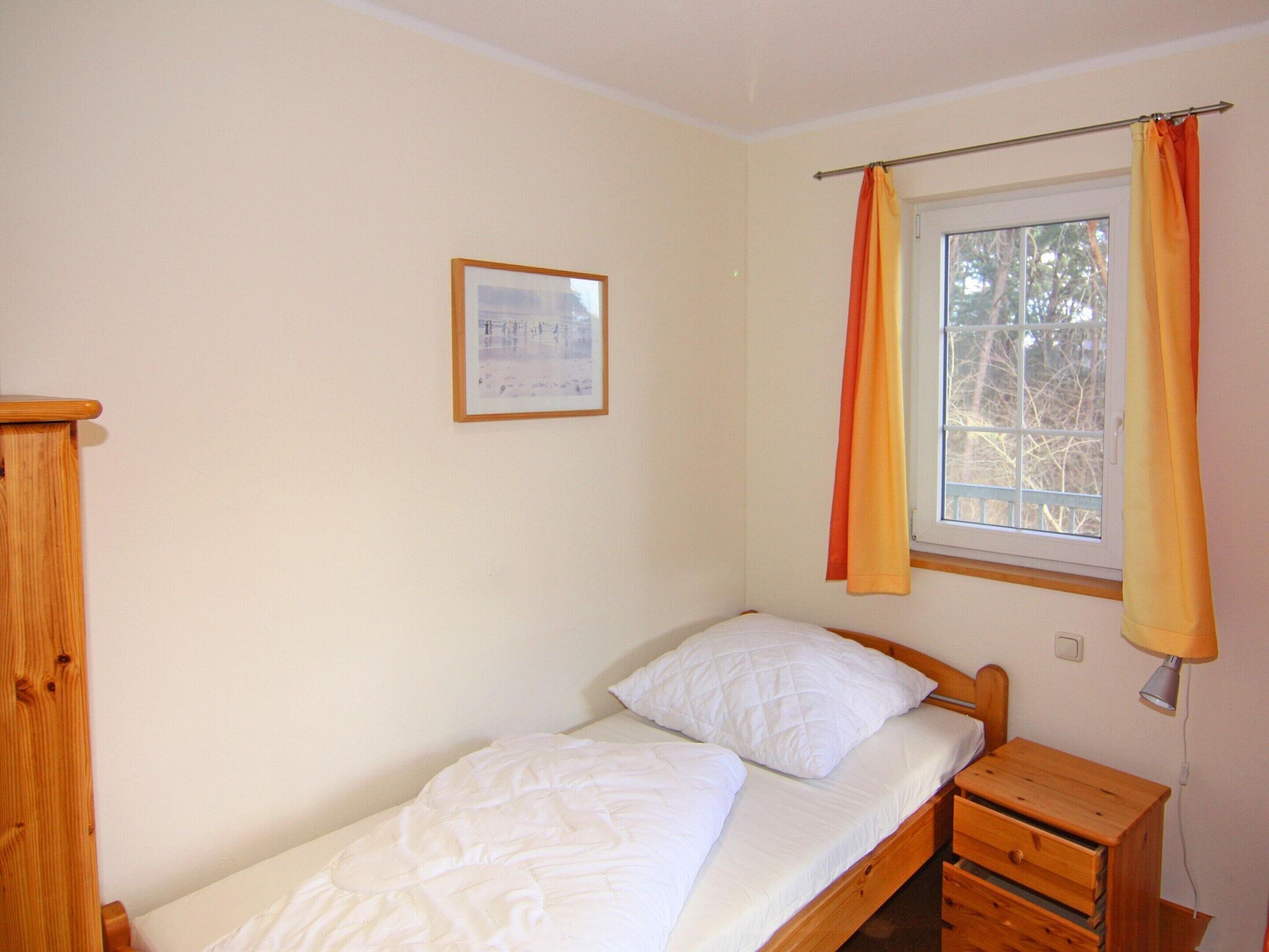 A small, single-bed room with a wooden bed frame, a nightstand, a bedside lamp, and a window with orange curtains.