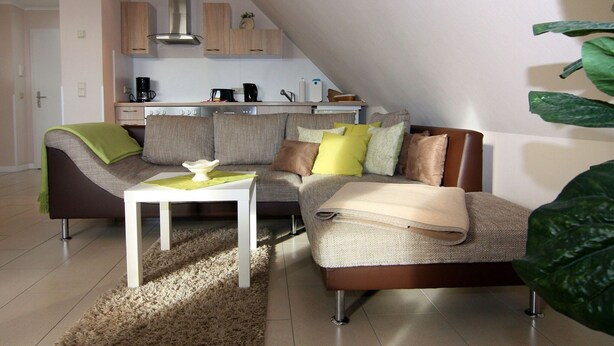 A modern living room with a sectional sofa, a coffee table, and a kitchen in the background. - Appartementhaus Strandloper, Zinnowitz (Zinnowitz)