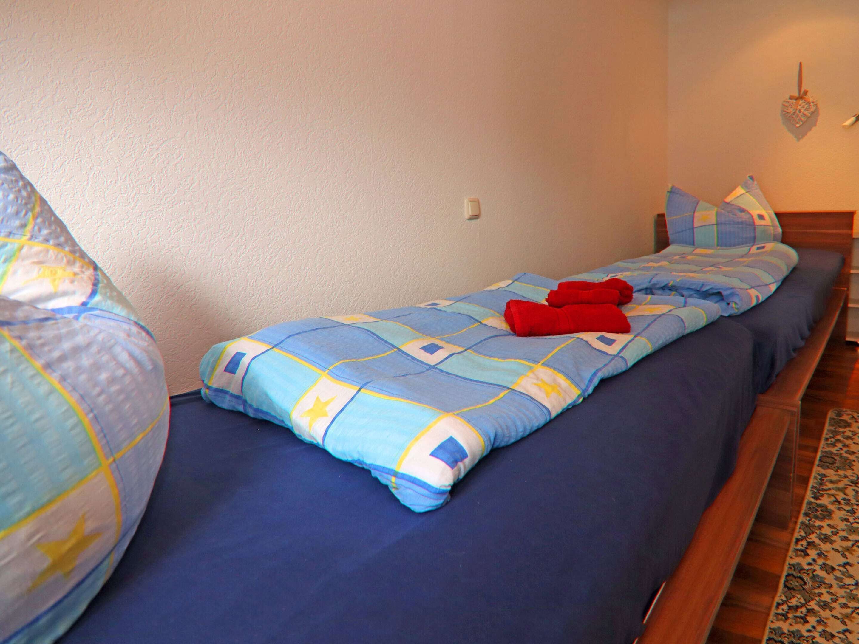 A single bed with a blue quilt and red towels, a wooden headboard, and a patterned rug.