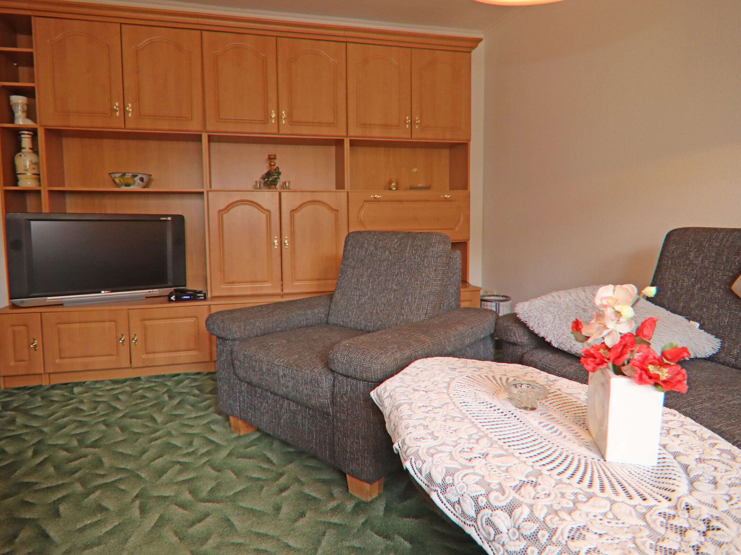 A living room with a television on a wooden cabinet, a grey sofa, and a table with a lace tablecloth and flowers.
